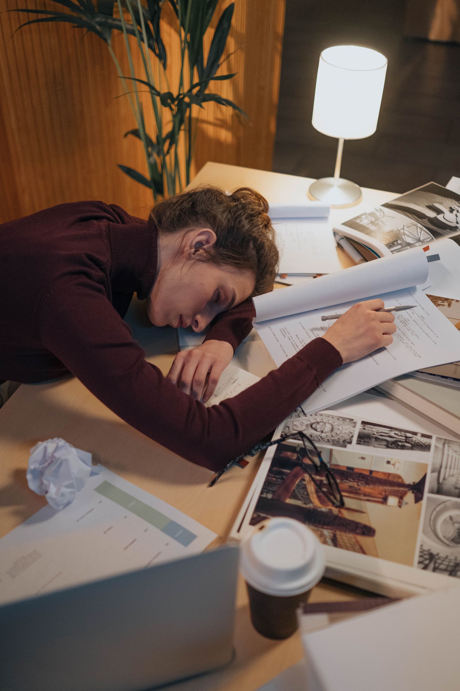 A person asleep at a cluttered desk late at night while the work waits on the page in front of them